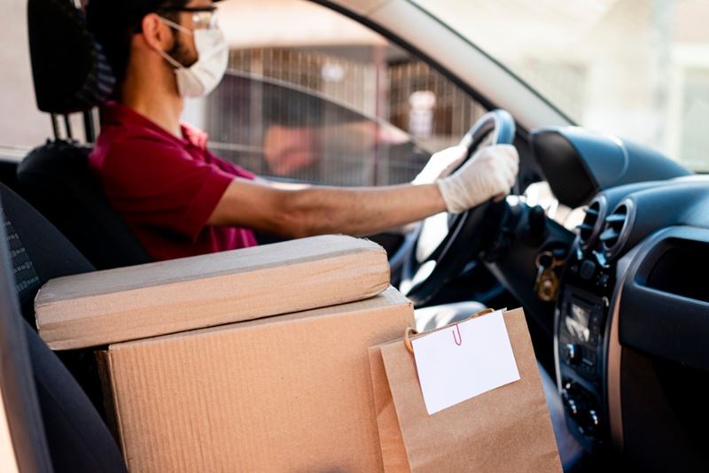 Hispanic male courier in a red shirt, face mask and gloves with orders in a car delivery service.